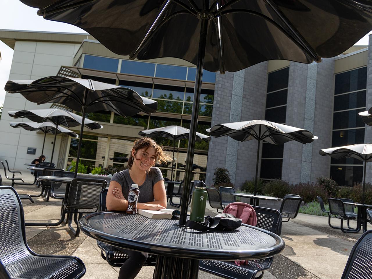 A students looks up from a book that she is reading at a table outside of the Reese Center.
