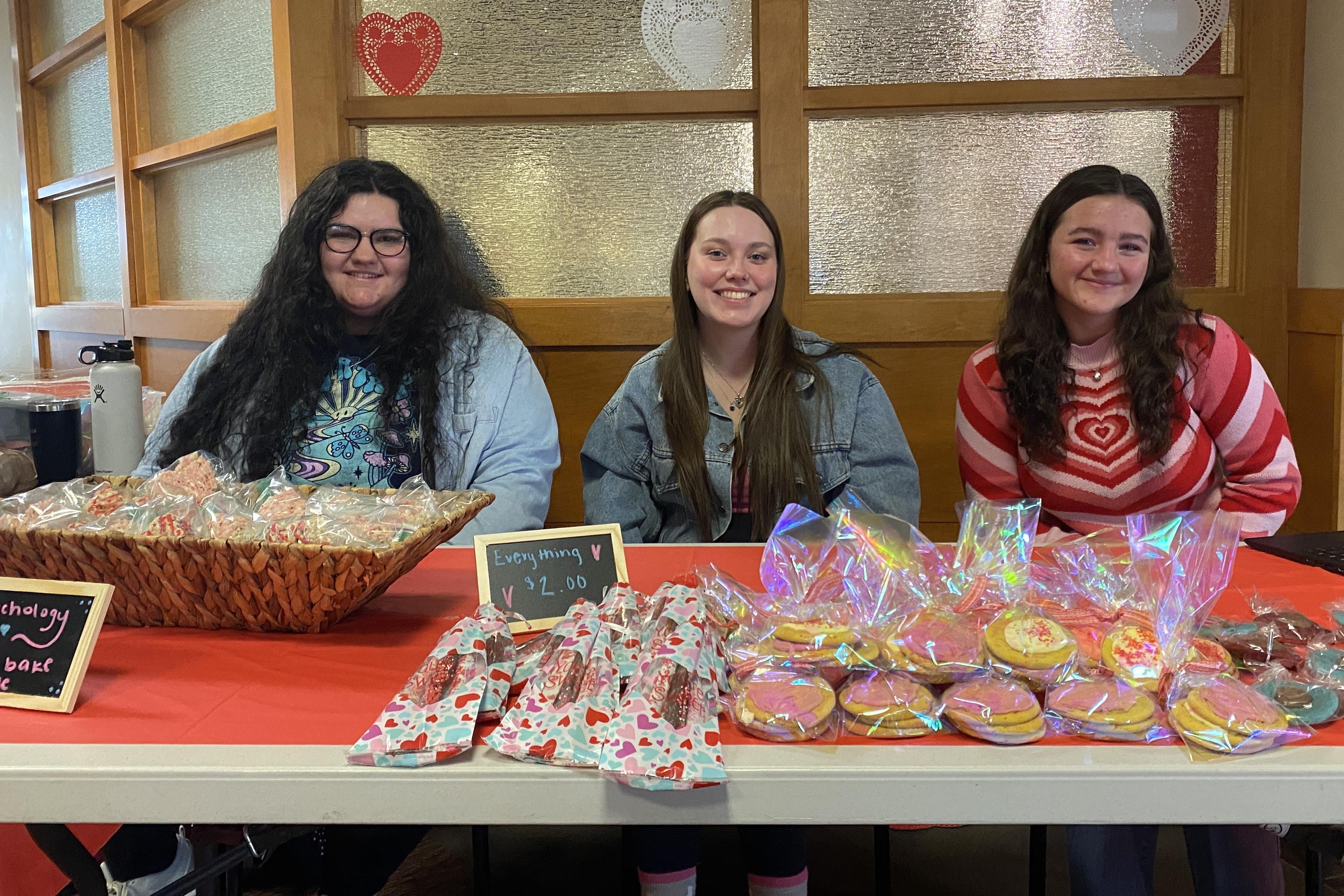 Students sit at a table topped with baked goods for sale.