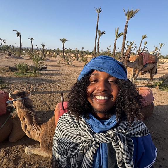 Student Ilyas Nur stands in a dessert in front of camels during an education abroad trip to Morocco.
