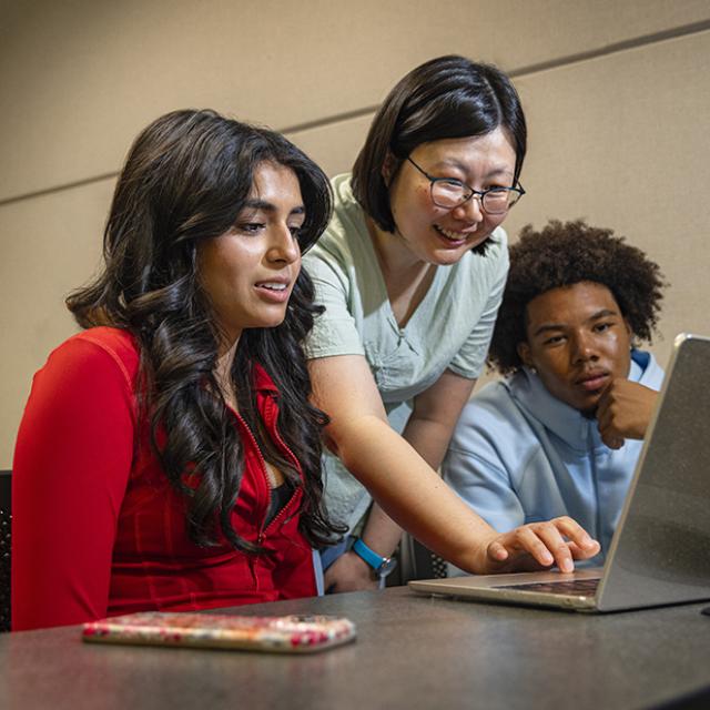 A professor stands behind two students seated at a table, reaching between them to use a laptop at which they are all looking.