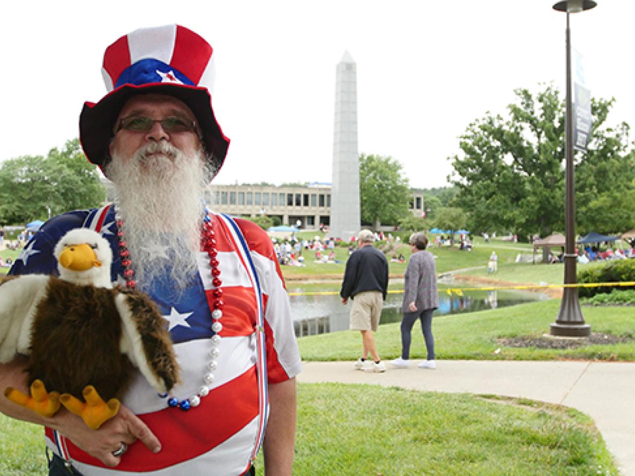 Man with toy stuffed eagle and red, white and blue clothing and hat smiling at the camera at July 3rd Fireworks and Concert