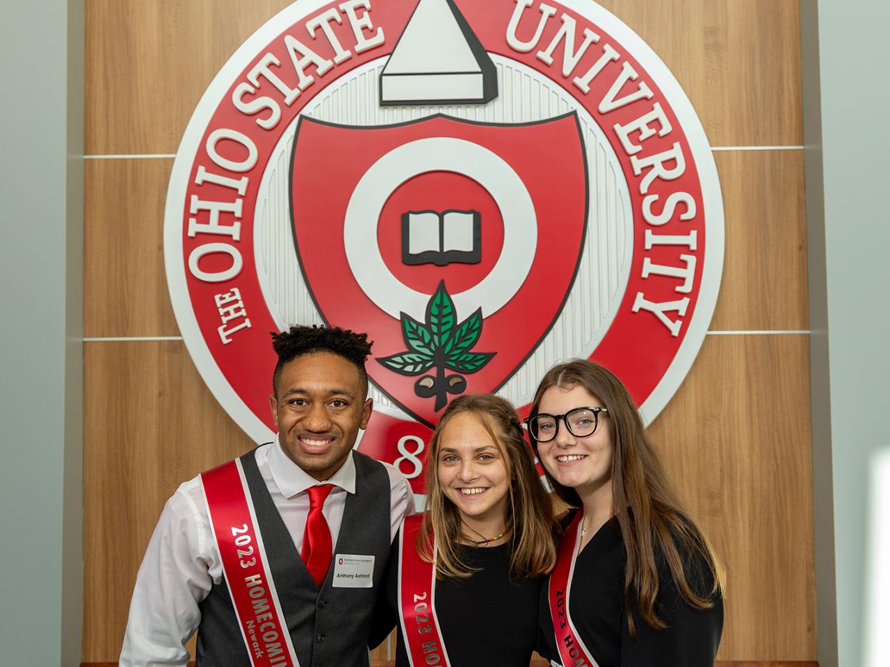 Three members of the Homecoming court stand in front of the university seal at the Longaberger Alumni House.
