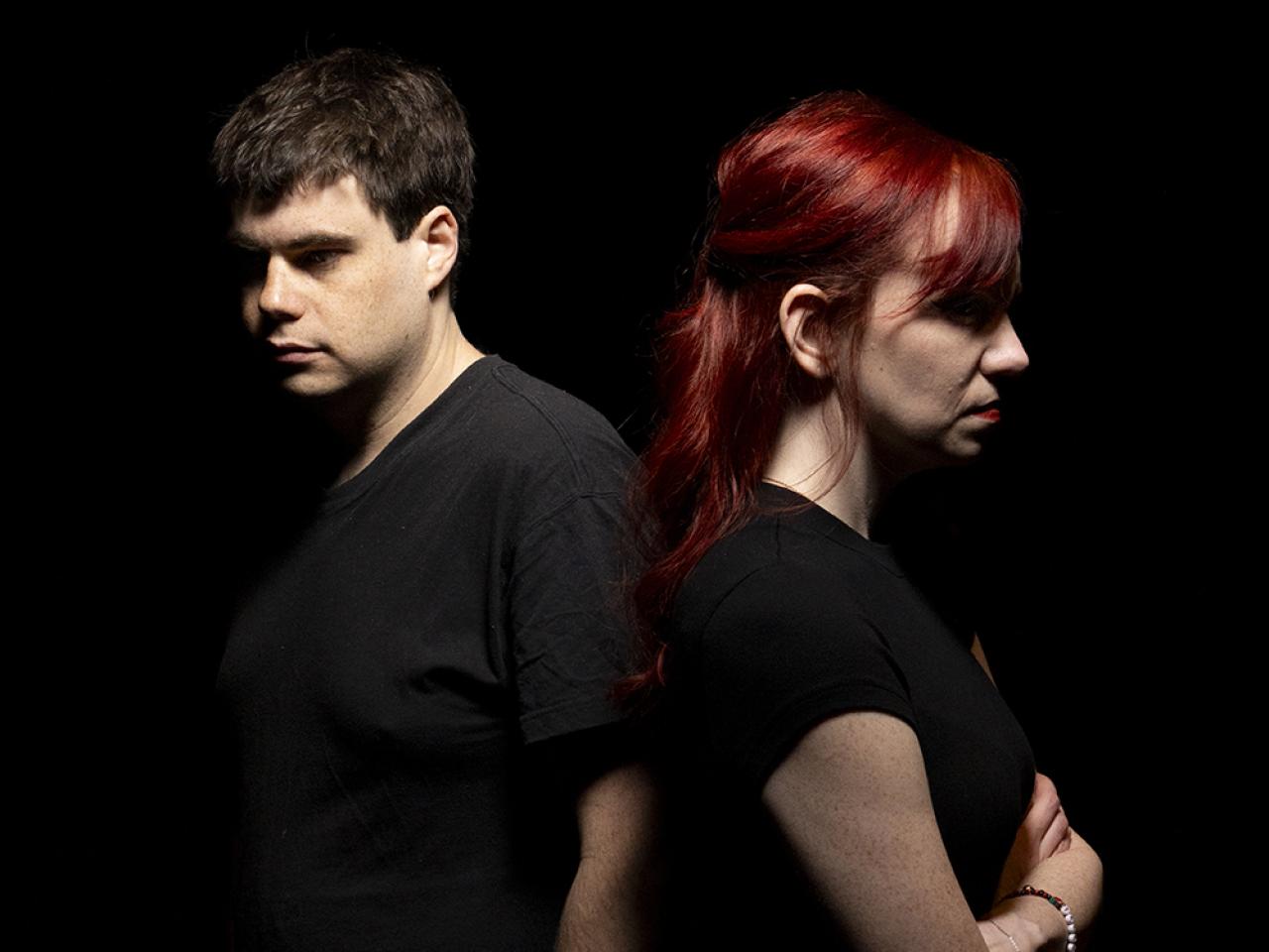 A man and woman stand back to back with unhappy expressions in a black space.