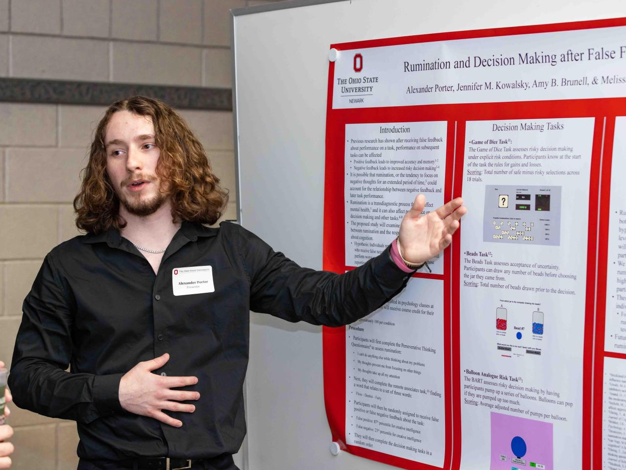 A student stands next to his poster presentation talking to a spectator and pointing at data on the poster.