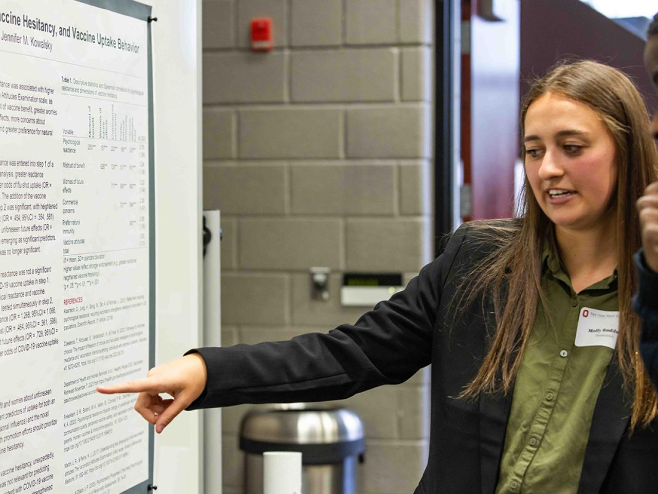 Student points to her research poster while talking to a bystander at the Student Research and Scholarship Forum
