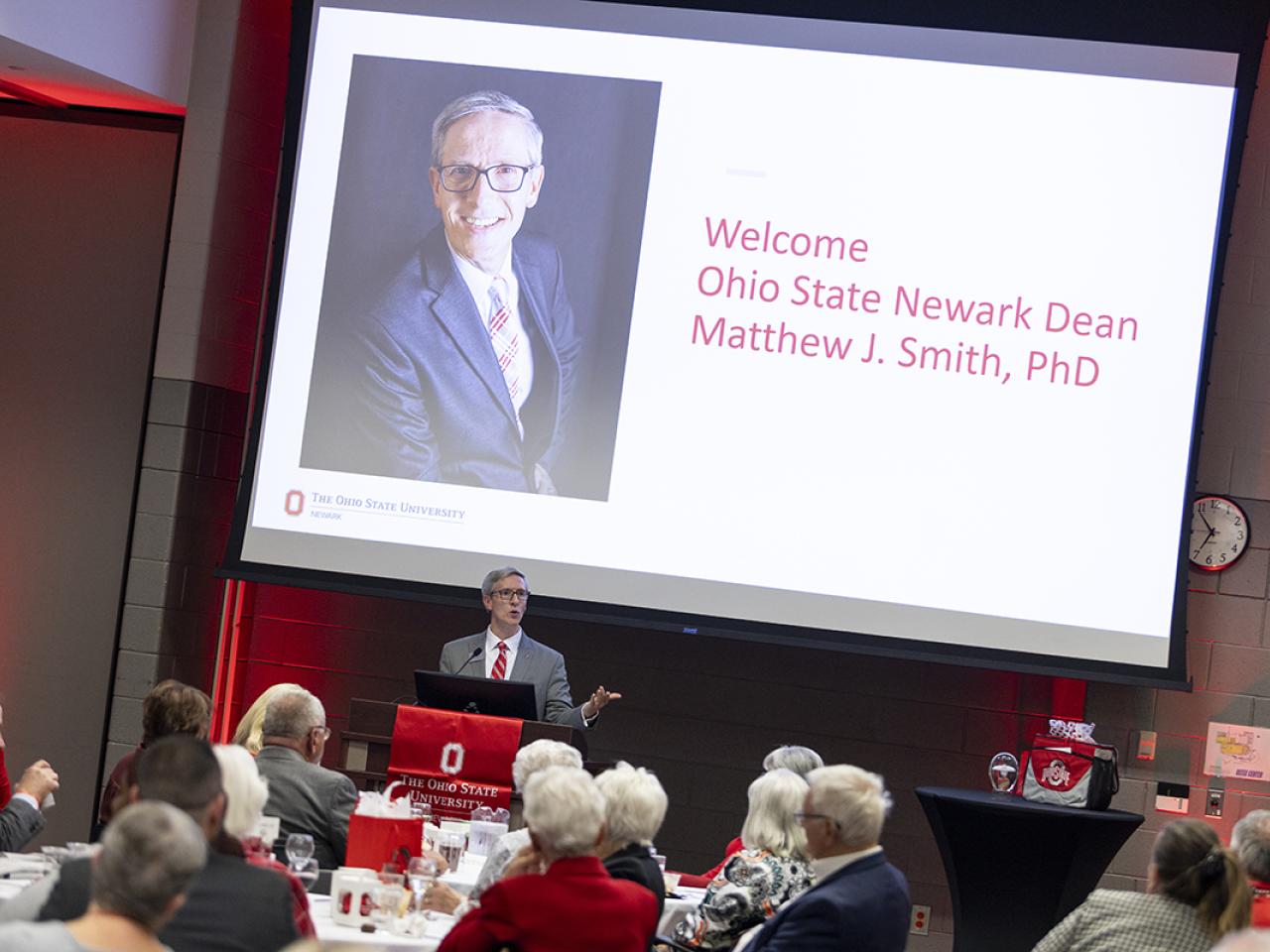 Dean and Director Matthew Smith stands at a podium addressing the crowd of alumni attending the Homecoming celebration.