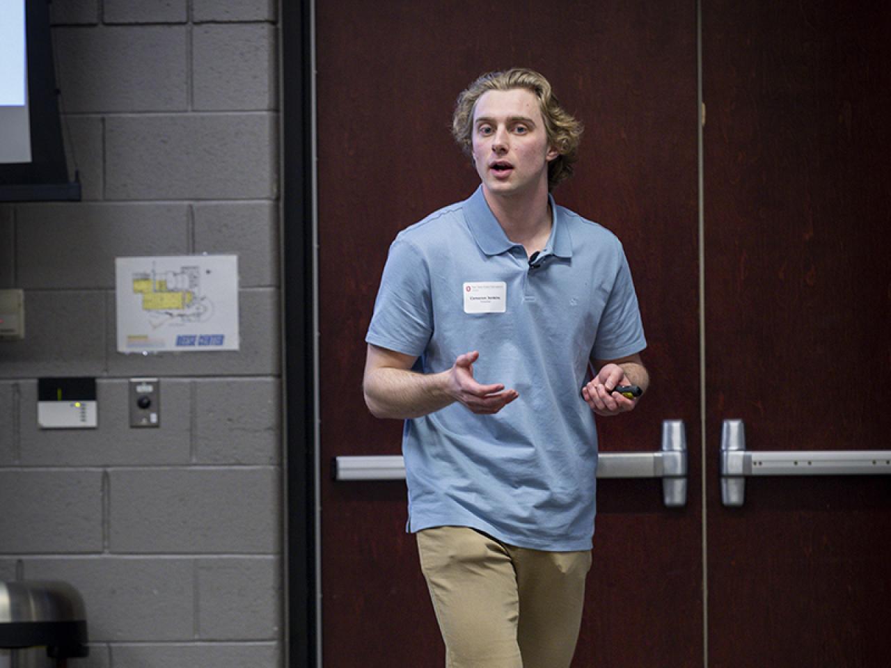 A student stands in the front of a room speaking about his research.