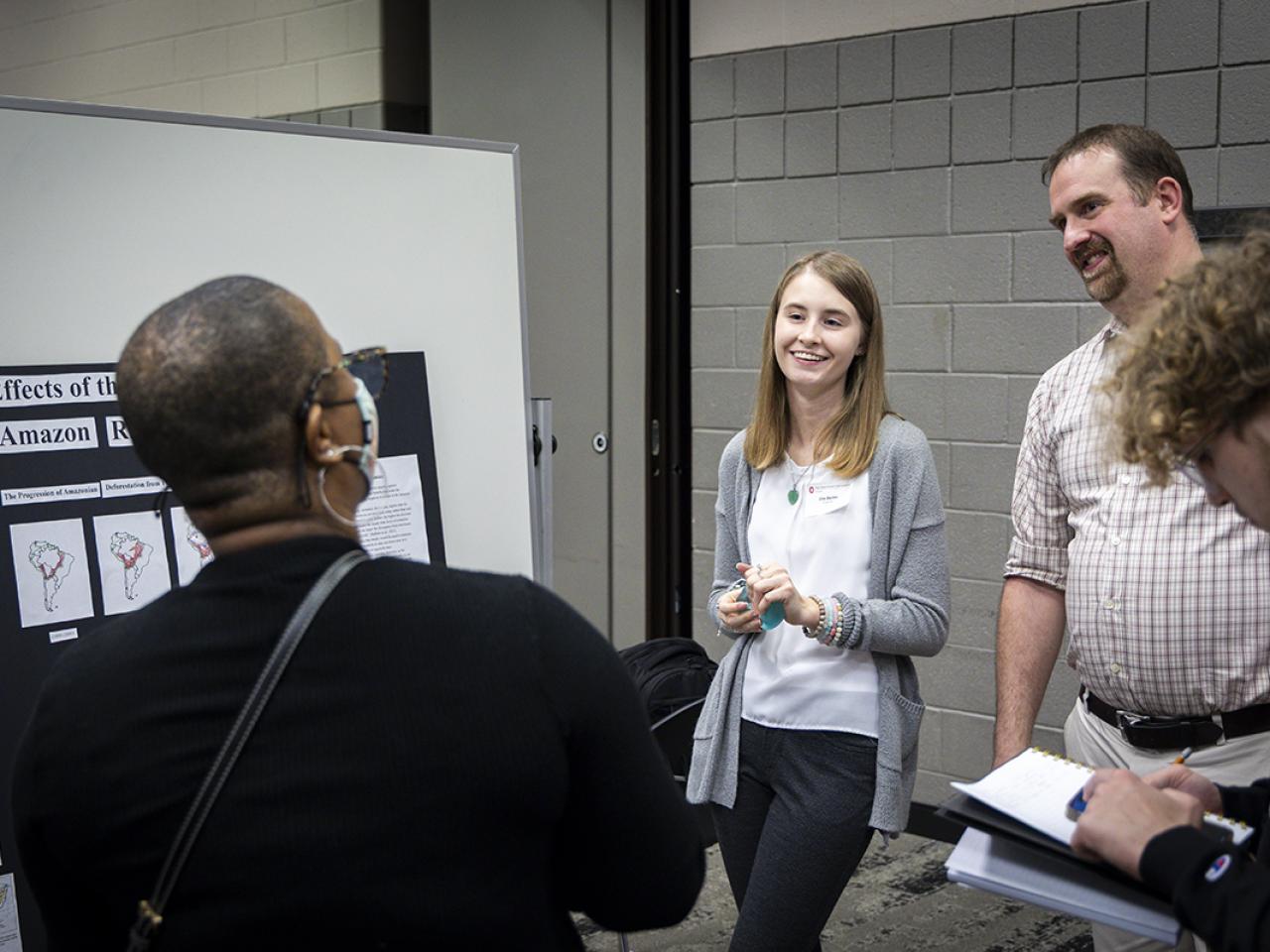 A student stands next to her poster presentation talking to a spectator. 
