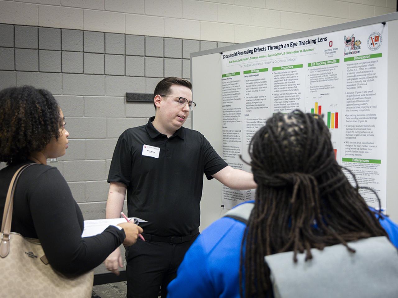 A student points to his poster while two onlookers watch.