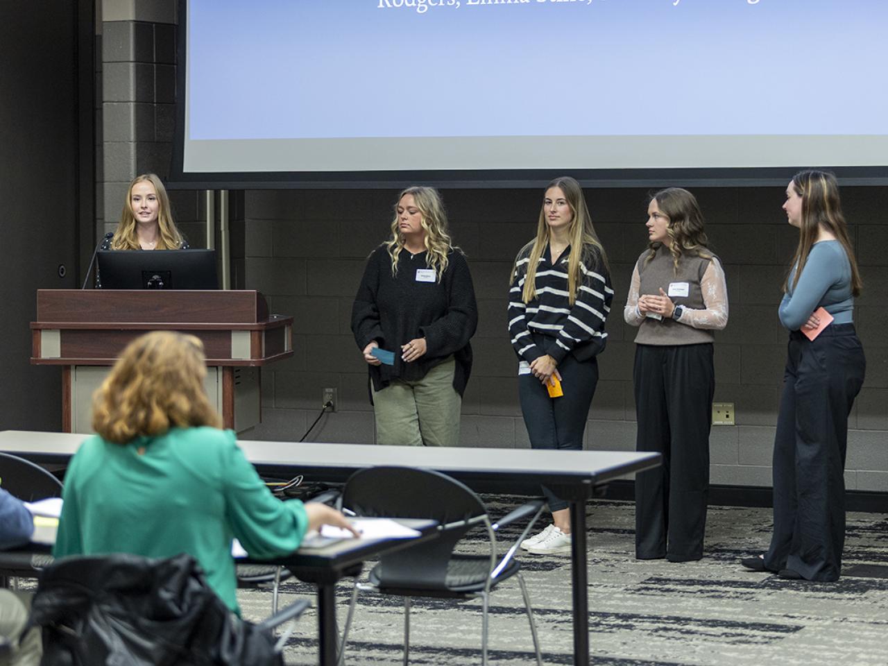 A student stands at a podium while four others stand at the front of the a crowd in a conference room.