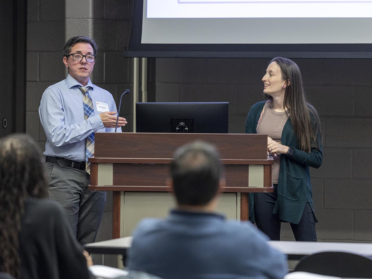 A student standing at a podium addresses a crowd while a second students looks at the first.