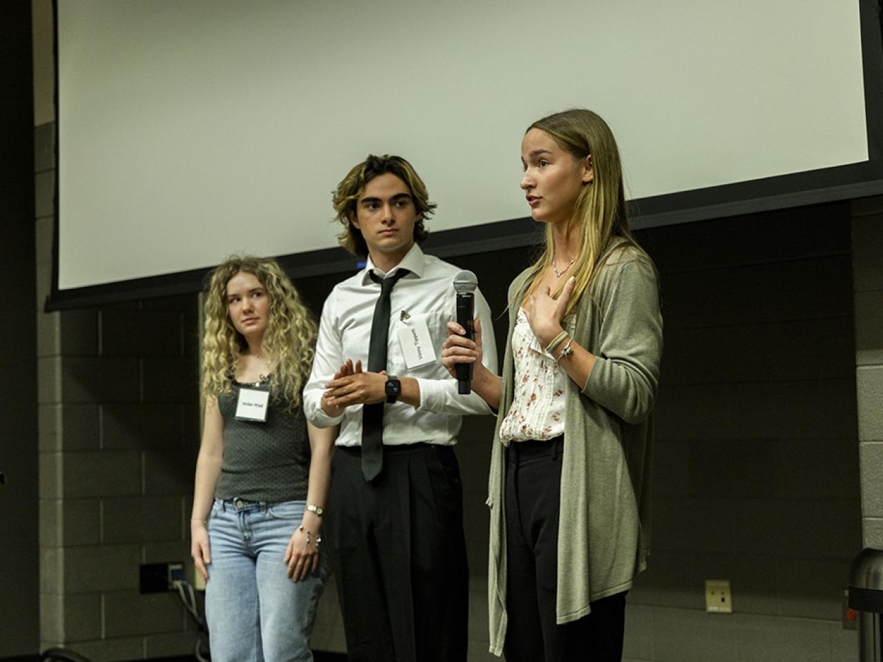 Carly Williams speaks into a microphone standing next to her co-presenters Jordan Waid and Vinny Tippett during their oral presentation at the Student Research and Creative Inquiry Forum.