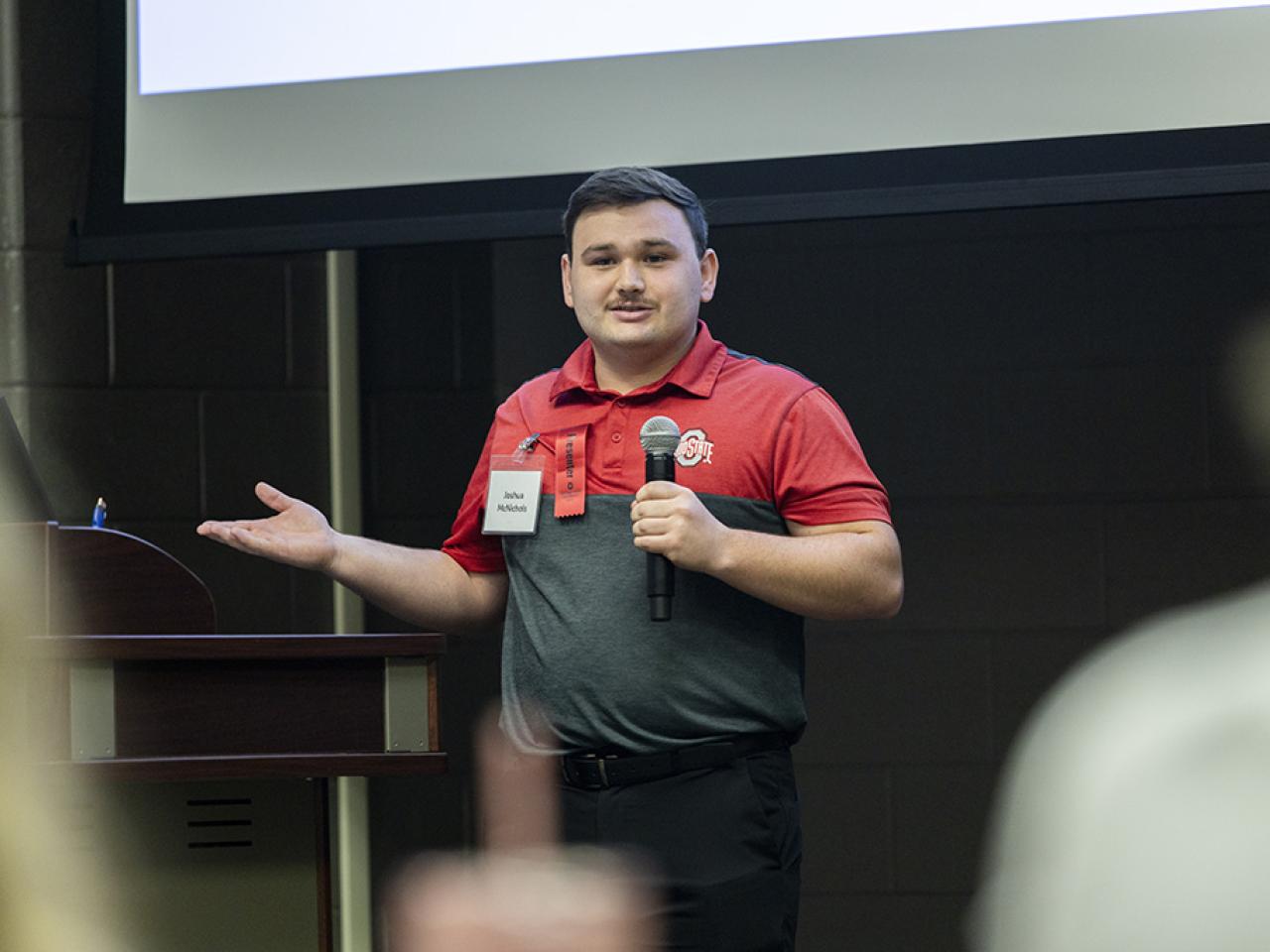  Joshua McNichols speaks into a microphone to an audience during his oral presentation at the Student Research and Creative Inquiry Forum