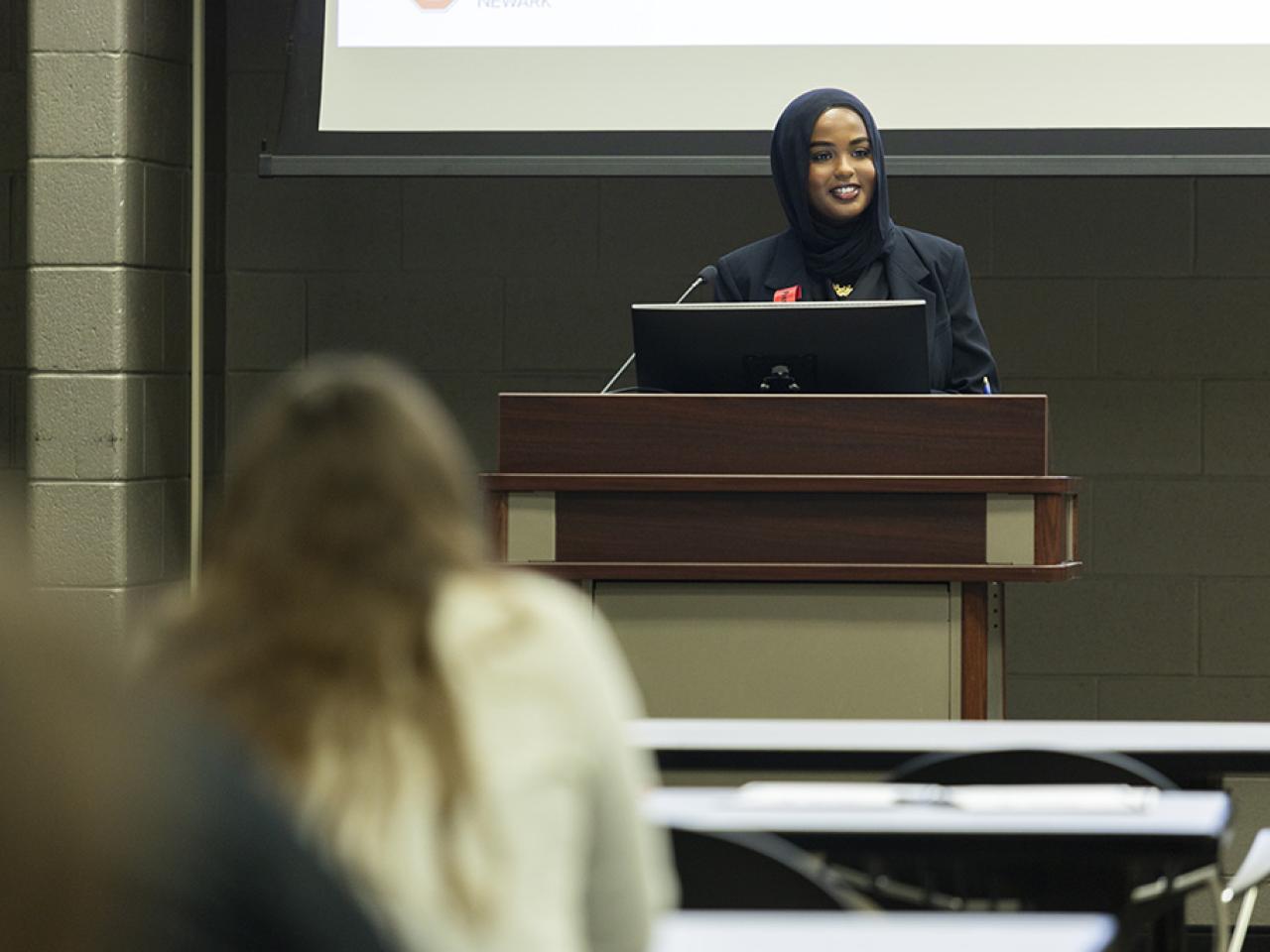 Nadia Faqi stands at a podium giving an oral presentation to spectators at the Student Research and Creative Inquiry Forum.