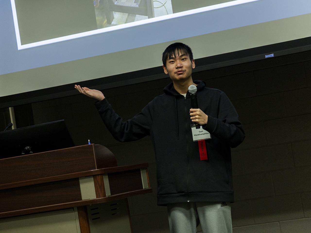 Sam Maokhamphiou stands with one handed lifted upward and the other holding a microphone during his oral presentation at the Student Research and Creative Inquiry Forum.