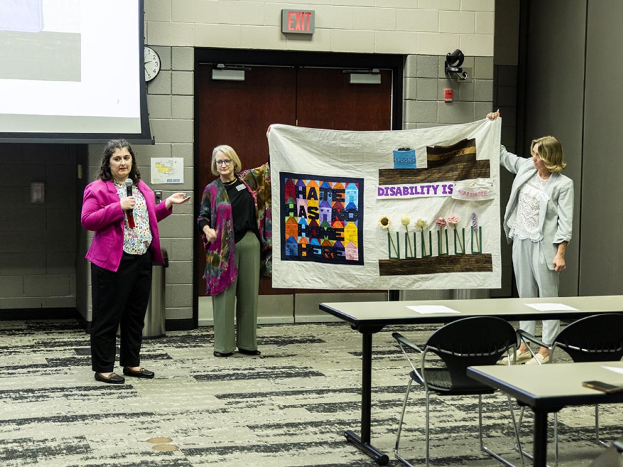 Sarah Ghiassi stands next to a quilted display speaking into a microphone during her oral presentation at the Student Research and Creative Inquiry Forum.