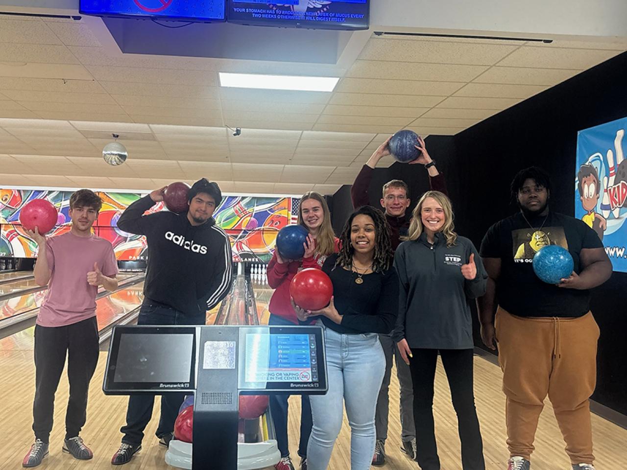 A group of students stand in line holding bowling balls in front of a bowling lane at a bowling alley. 