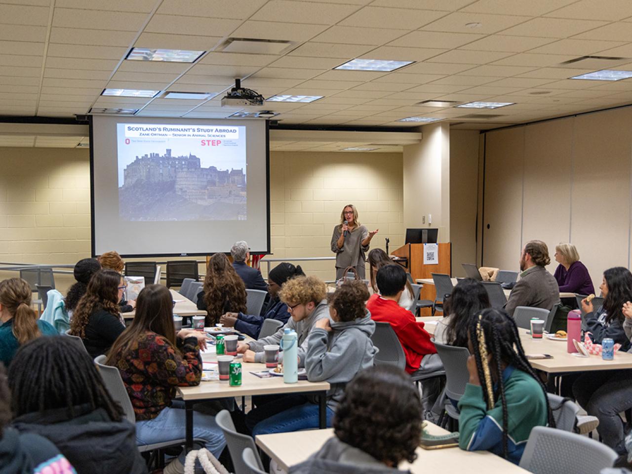 A presenter stands delivering a speech in front of a room full of students.