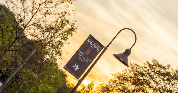A flagpole bearing a flag that says, "Welcome to Ohio State Newark" on campus with sunrise in the background.