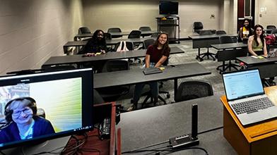 Students sit in a classroom with a professor on a computer.