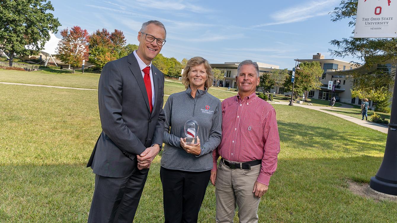 Kelly Lyons holds a glass trophy while standing between Mark Lyons and Dean William L. MacDonald on the Newark campus.