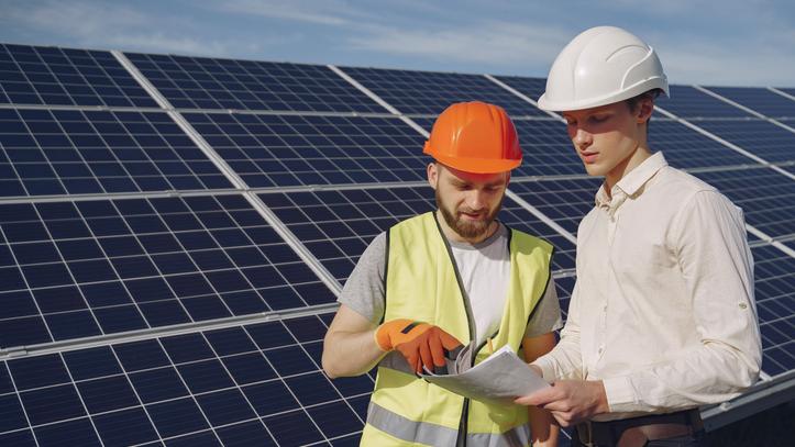 A man in a business attire and a man in a safety vest and gloves look at a paper while standing in a solar panel field.