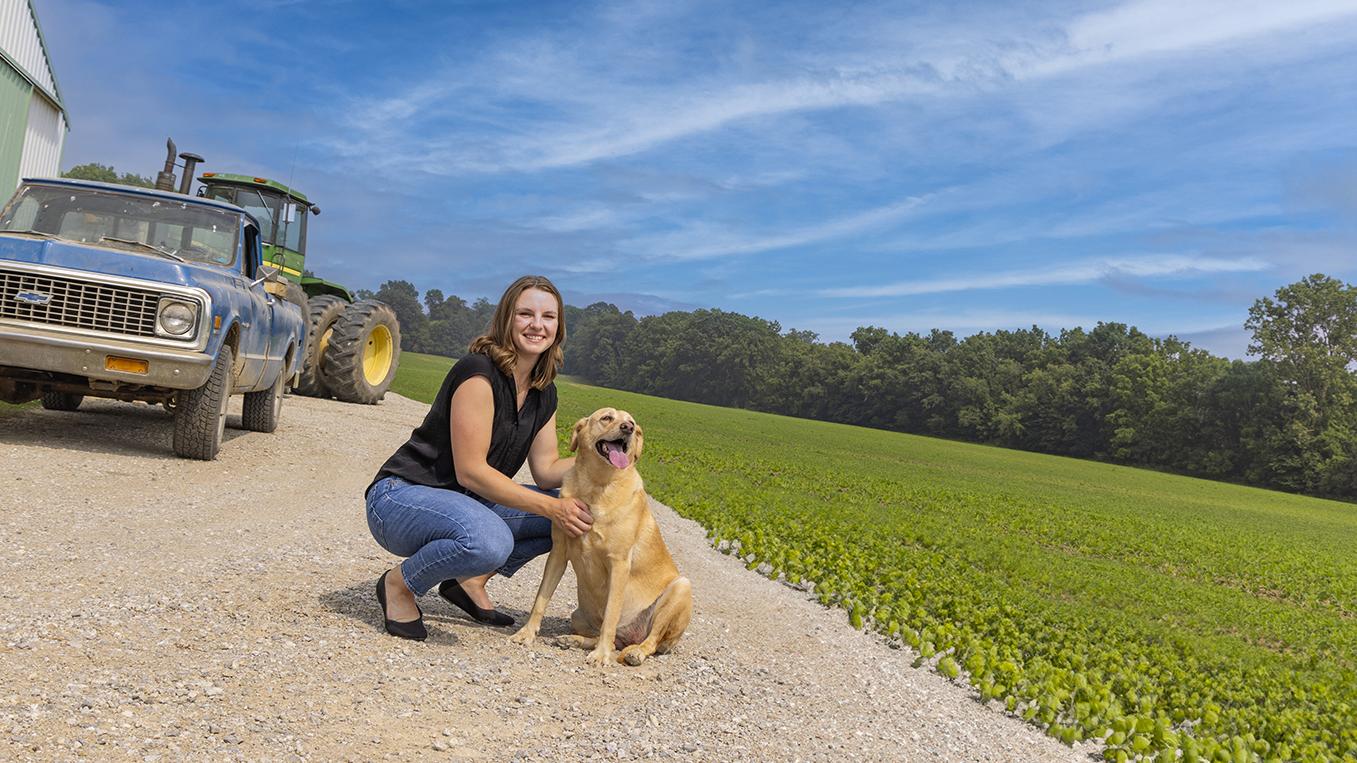 Madi Layman poses with a dog in front of a barn and field of soybeans.