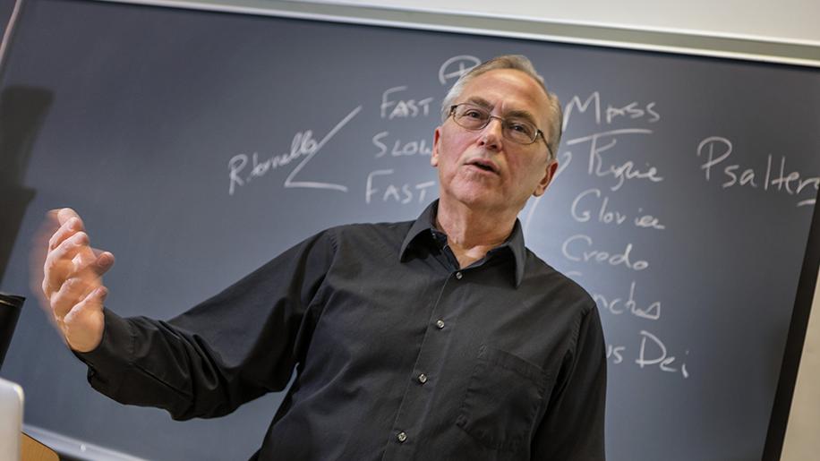 Paul Sanders stands in front of a chalk board motioning with his hand to an unseen class.