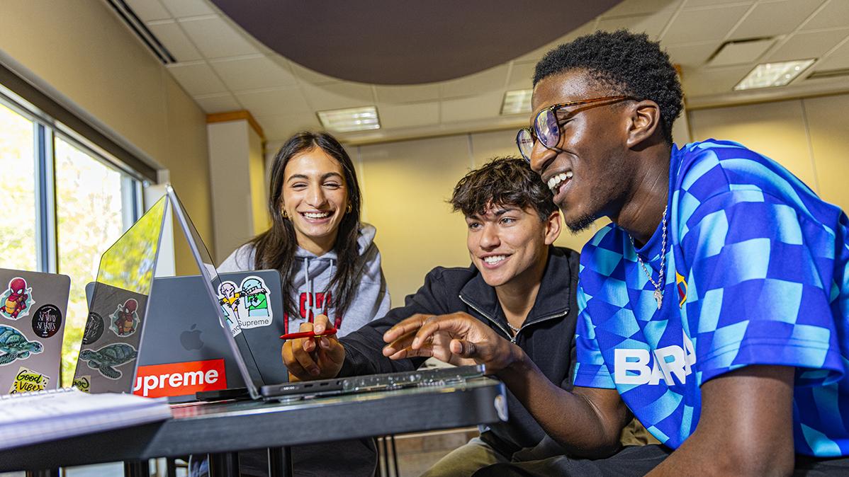 Three students sit around a table looking at a laptop and smiling. 