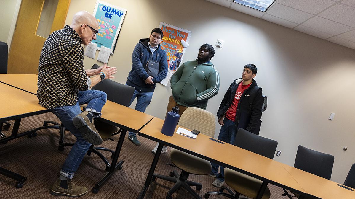 A professors sits on a desk speaking to three students who are standing around him in a classroom.