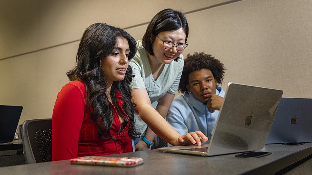 A professor stands behind two students seated at a table, reaching between them to use a laptop at which they are all looking. 
