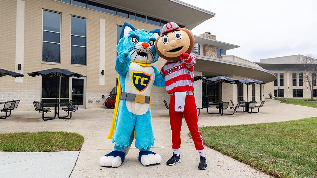 TC the Central Ohio Technical College cat mascot and Brutus Buckeye stand with one arm around each other and the other held out to point a finger toward the viewer.