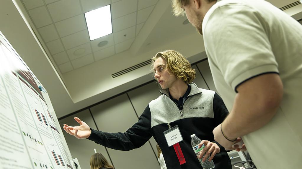 Student Cameron Jenkins raises his hand and looks at his poster presentation as a bystander listens.