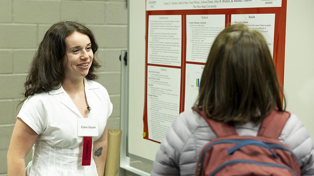 Claire Gaulin stands next to her poster presentation speaking to an attendee at the Student Research and Creative Inquiry Forum.