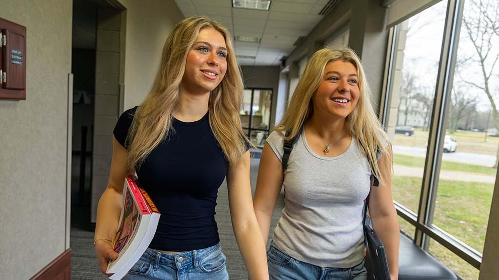 Sisters Avery and Reese Staten walk down a hallway carrying books and a laptop looking ahead with bright eyes and with smiles.