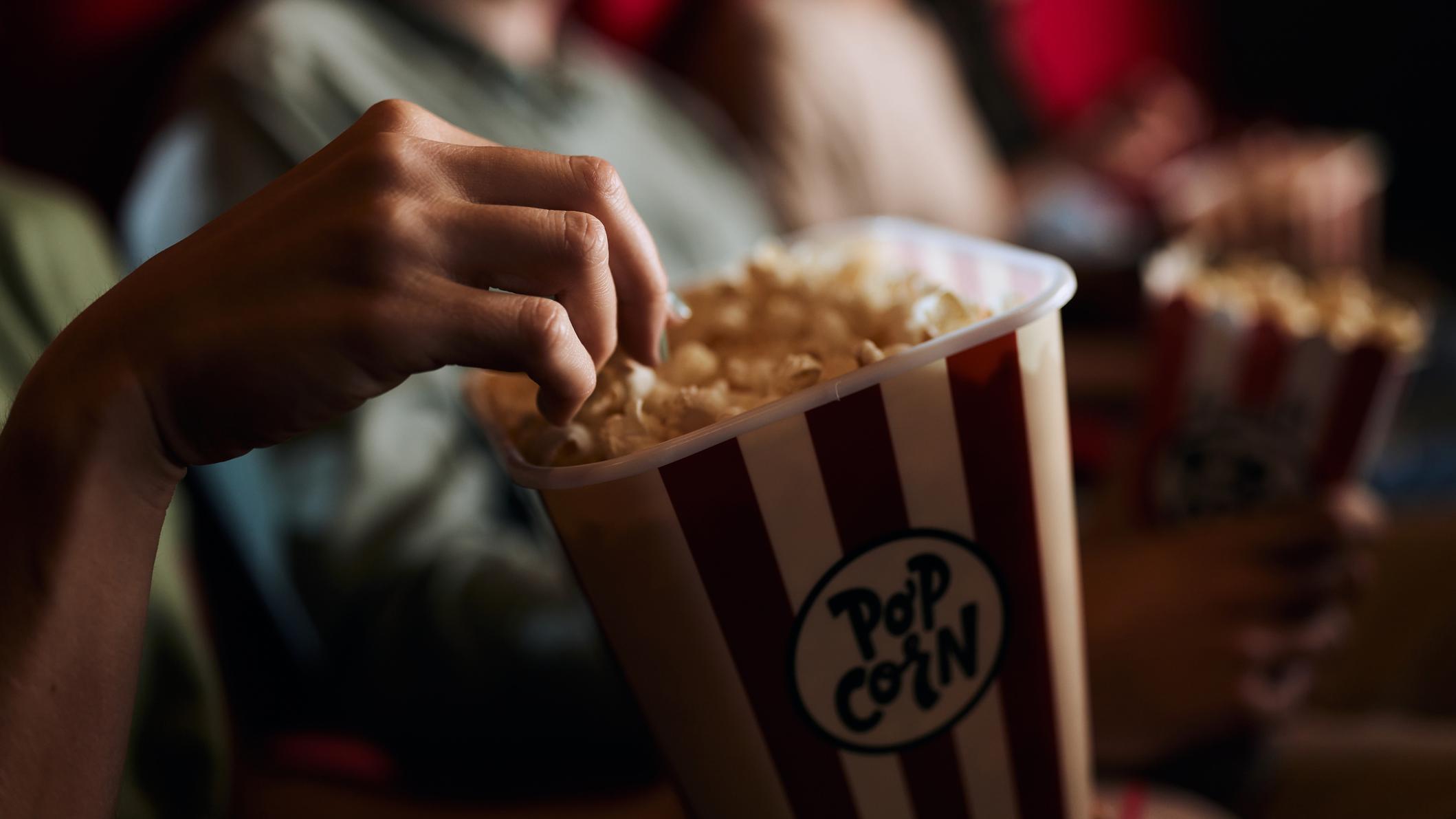 A hand reaches into a bucket of popcorn in a theater.