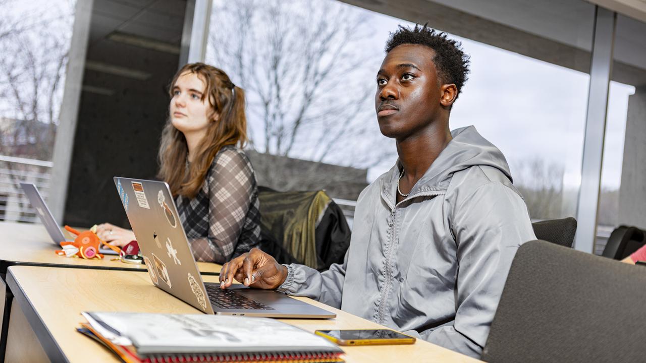 Two students sit side-by-side at a desk with open laptops looking up to the front of the classroom