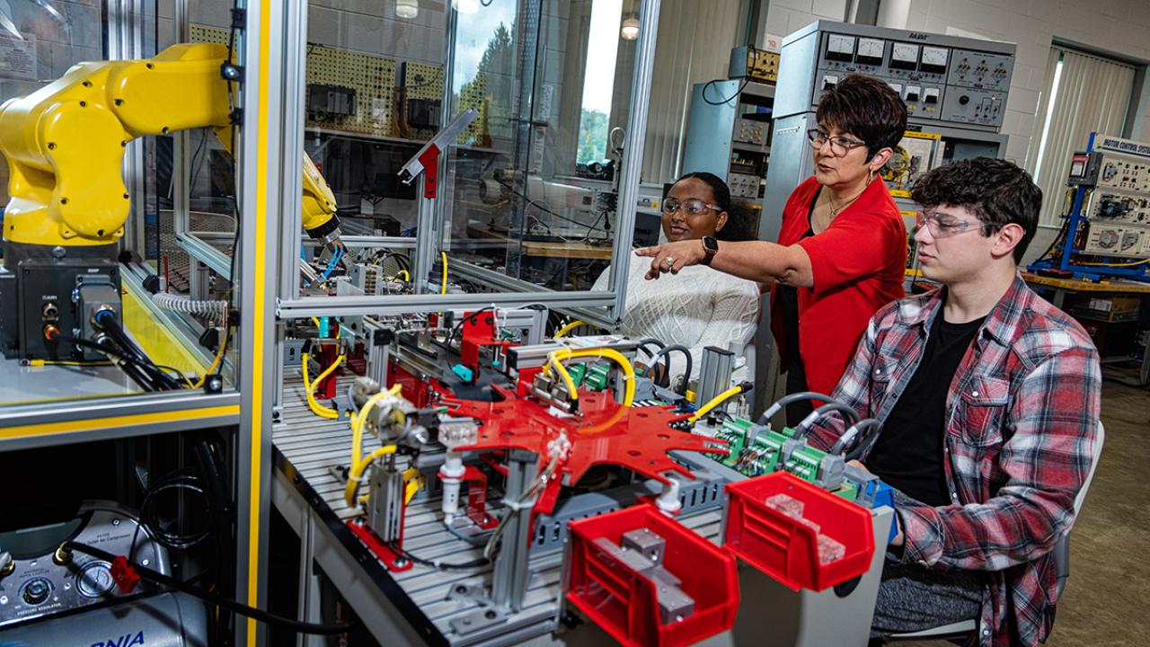 Engineering professor stands between students who are seated at engineering equipment. 