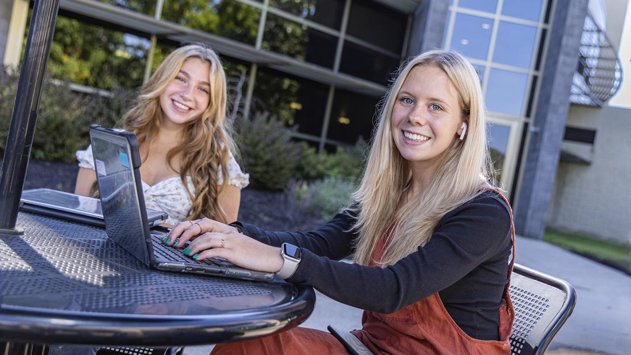 A students sits at an outdoor table with her hands on an open laptop smiling at the camera and with a friend smiling next to her.