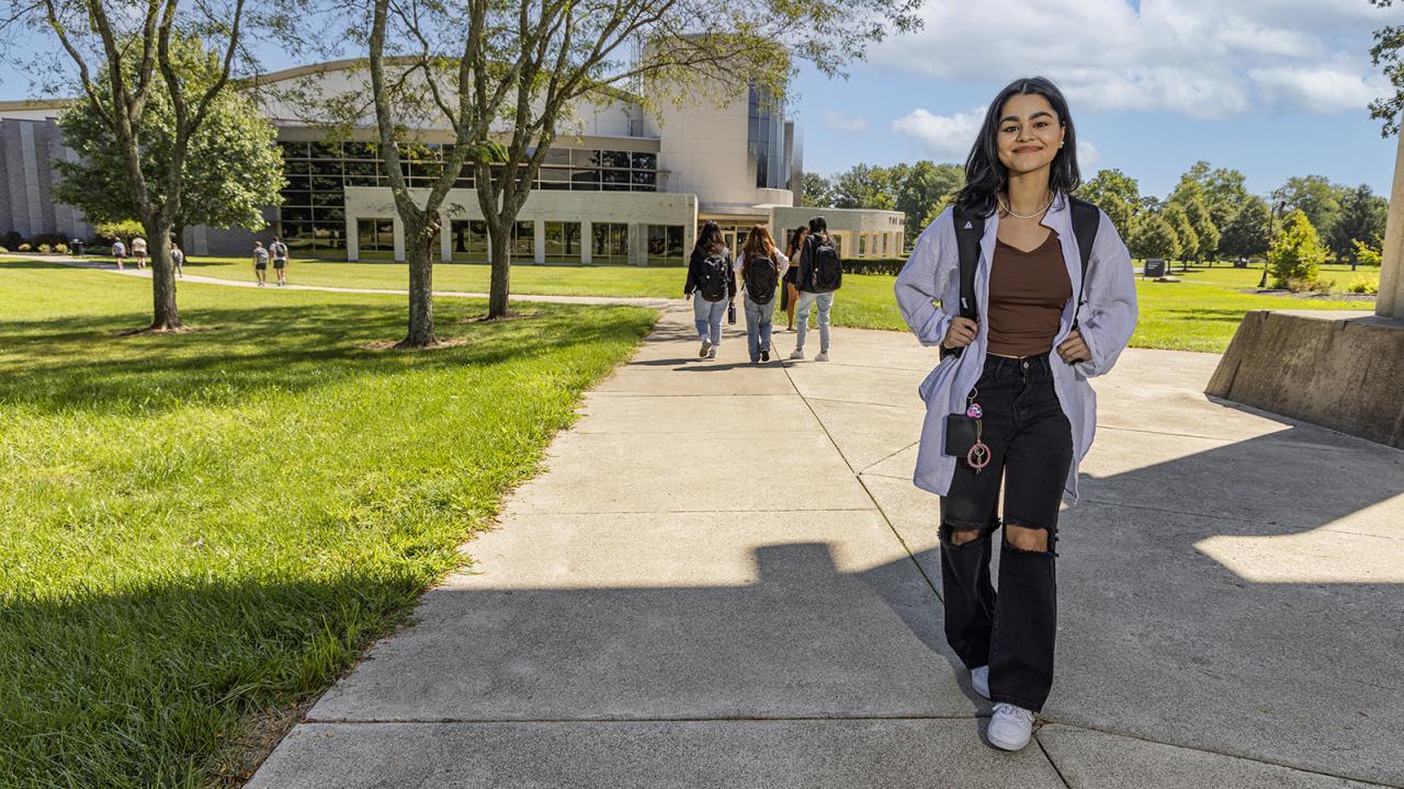 A female student walks toward the camera smiling outside of the classroom building. 