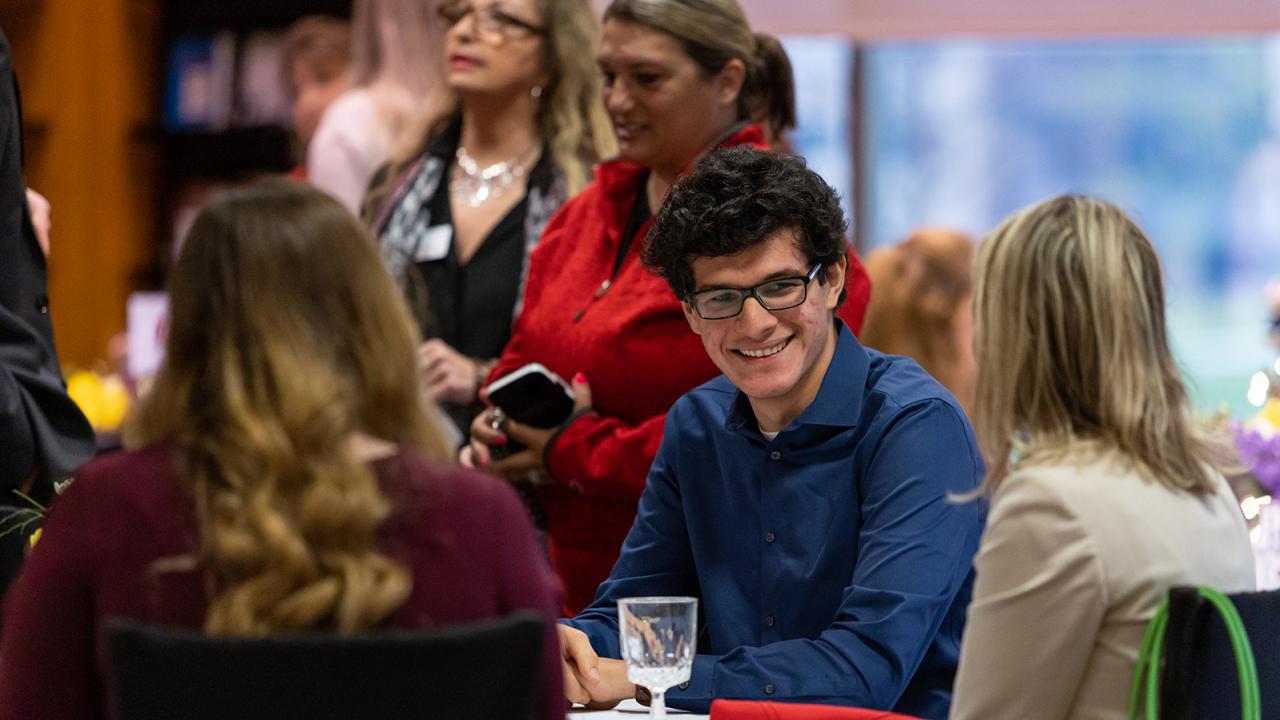 A male student sits at a table conversing with an alumna in a crowded room during the Dinner for 12 Buckeyes event.