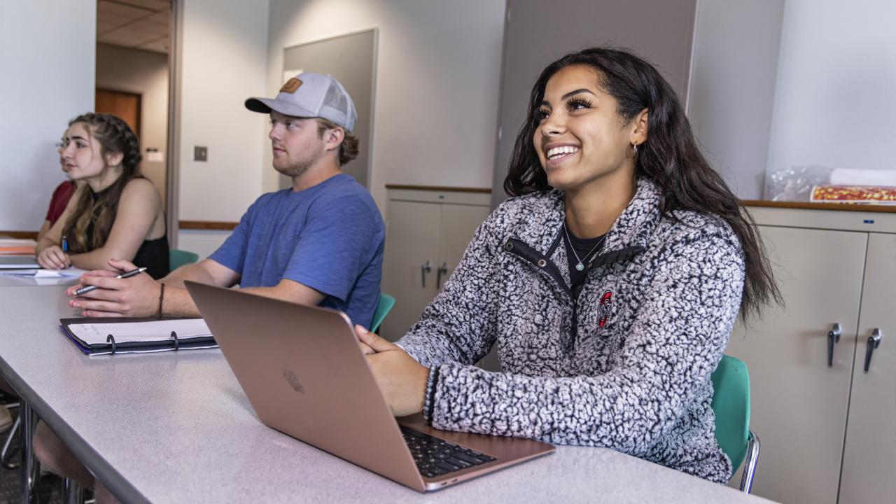 A student sits at a desk with other students in front of a laptop looking ahead with a smile 