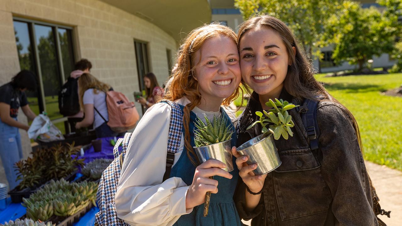Two female students stand together holding potted plants.