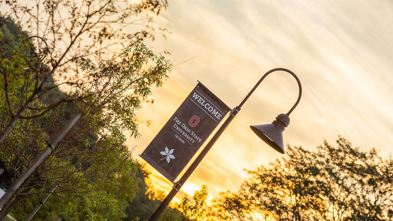 A flagpole bearing a flag that says, "Welcome to Ohio State Newark" on campus with sunrise in the background.