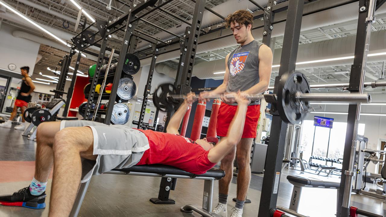 A student uses a bench press while another spots him. 