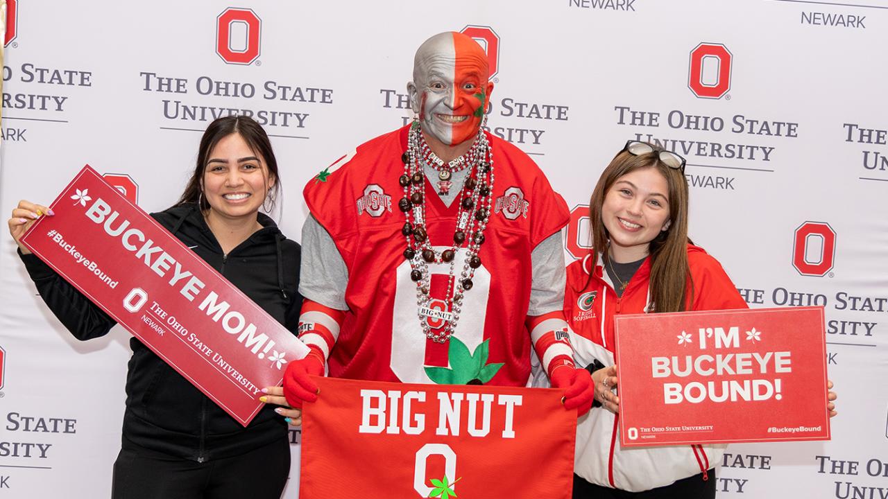 A woman holds a "Buckeye mom" sign and a student holds a "Buckeye bound" sign as they pose with the Ohio State super fan "Big Nut."
