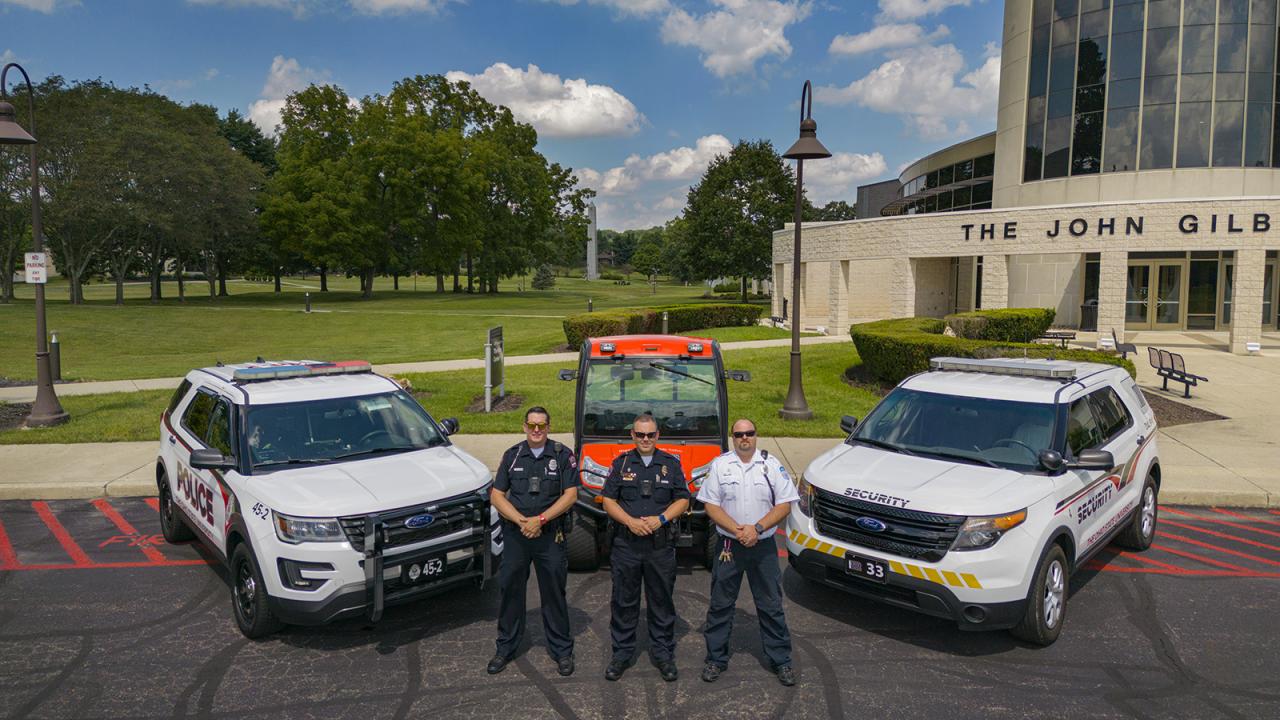 Two police officers and a security guard stand in front of police vehicles on campus.