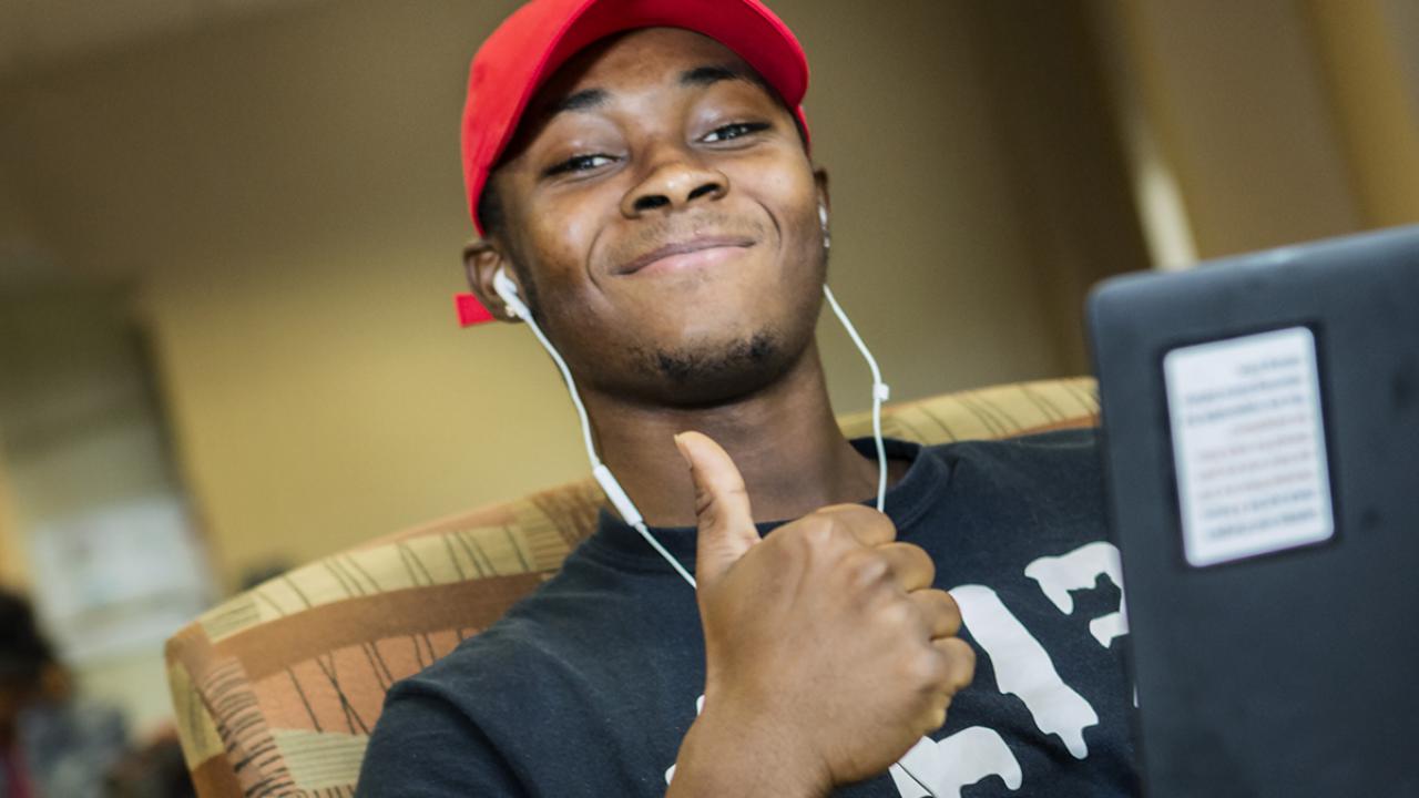 A student seated with a laptop on his lap gives a smile and thumbs up.