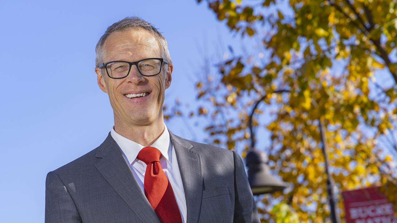 Portrait of Dean/Director William MacDonald on campus McConnell Hall in the background and yellow leaves on the trees in the fall.