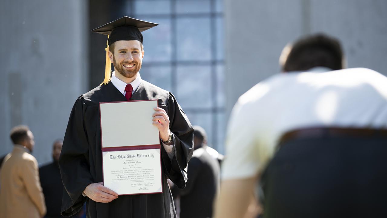 A male graduate dressed in academic regalia holds up his diploma at commencement.